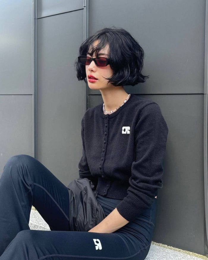 Asian model in streetwear look with black bob haircut, sunglasses and cropped cardigan against grey wall