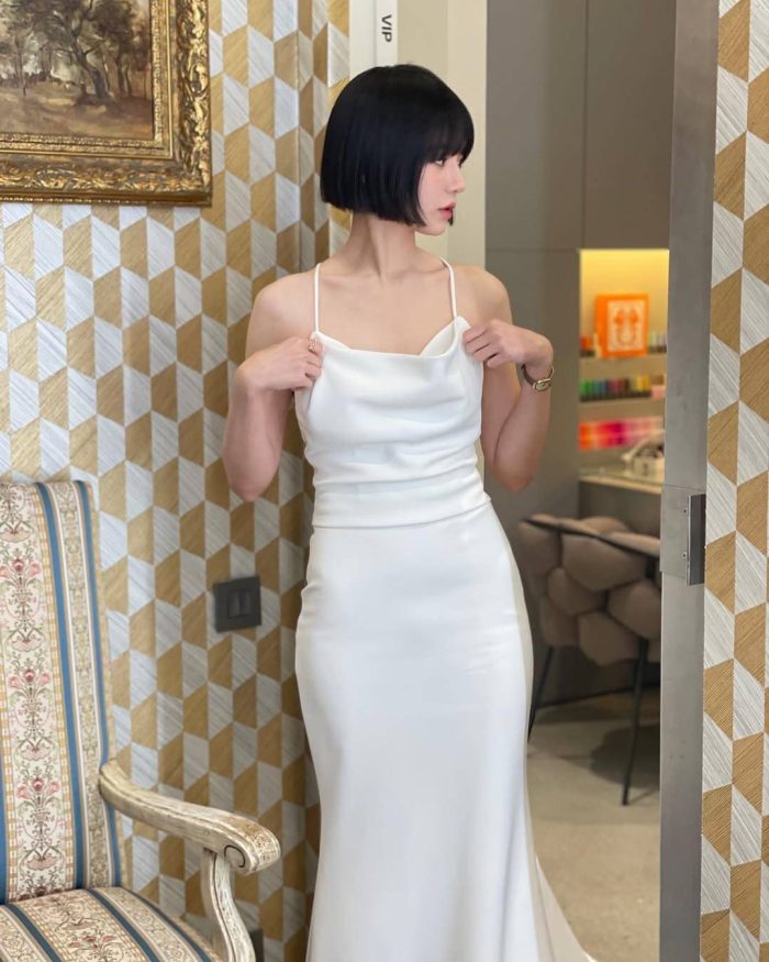 Model in elegant white silk slip dress with cowl neck and fitted silhouette against geometric wallpaper