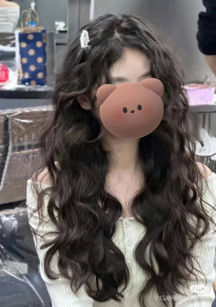 Woman with long curly dark hair wearing white sweater and hair clip with bear emoji covering face