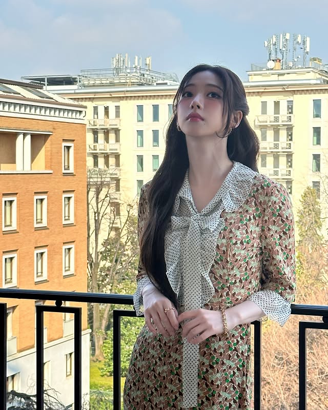 Asian woman with long black hair in floral dress on balcony overlooking city buildings