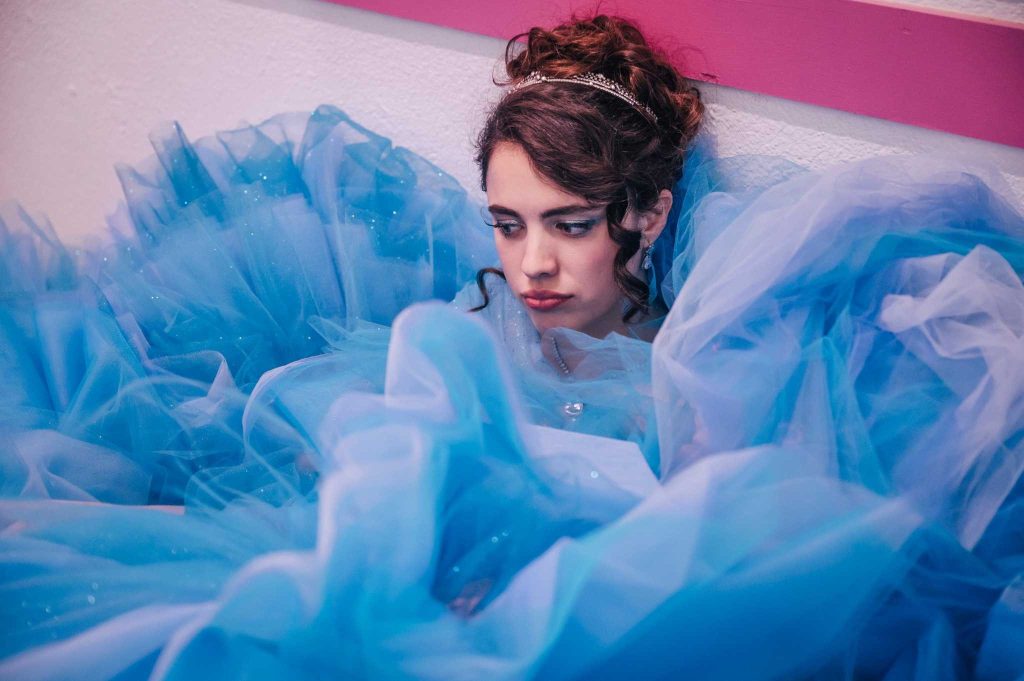 Person in elegant updo and tiara surrounded by flowing light blue tulle fabric in dreamy fashion portrait