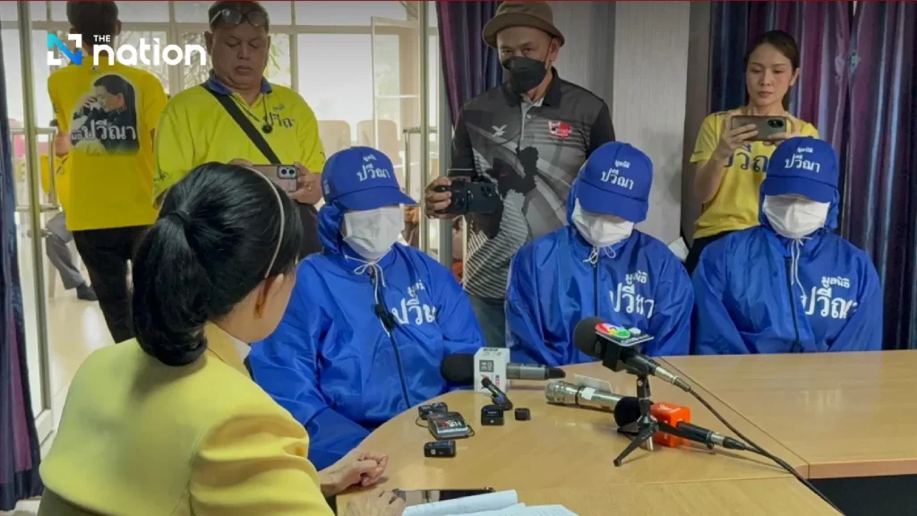Press conference with three people in blue uniforms and caps seated at table with microphones, while others in yellow attire observe