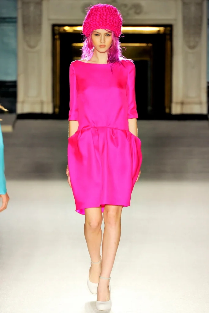 Model wearing vibrant hot pink dress with matching knit beanie walking runway at fashion show