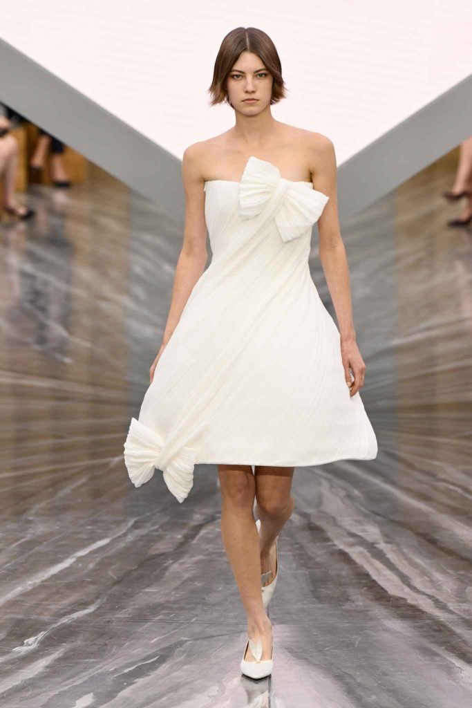 Model in dramatic black V-neck column dress with structured shoulders and cream opera gloves, wearing ornate crystal-embellished mask and walking on hardwood runway with string lights and potted plants backdrop.
