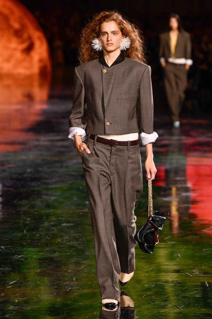 Model in cropped checked suit with rolled sleeves, white undershirt, and wide-leg trousers, wearing large white feather earrings and carrying black handbag with gold chain on reflective runway with warm lighting bac
