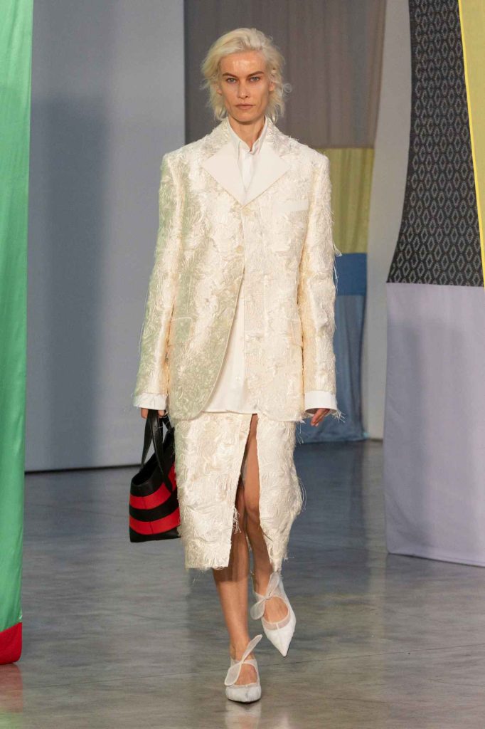 Model with platinum blonde hair wearing textured ivory suit ensemble featuring brocade blazer and midi skirt with front slit, paired with white ballet flats and striped tote bag against colorful backdrop