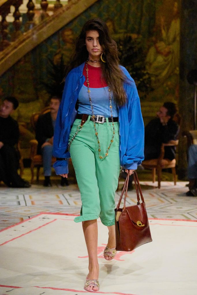 Model in color-block outfit with red top, blue cardigan, mint green capris, and brown leather tote bag on ornate runway