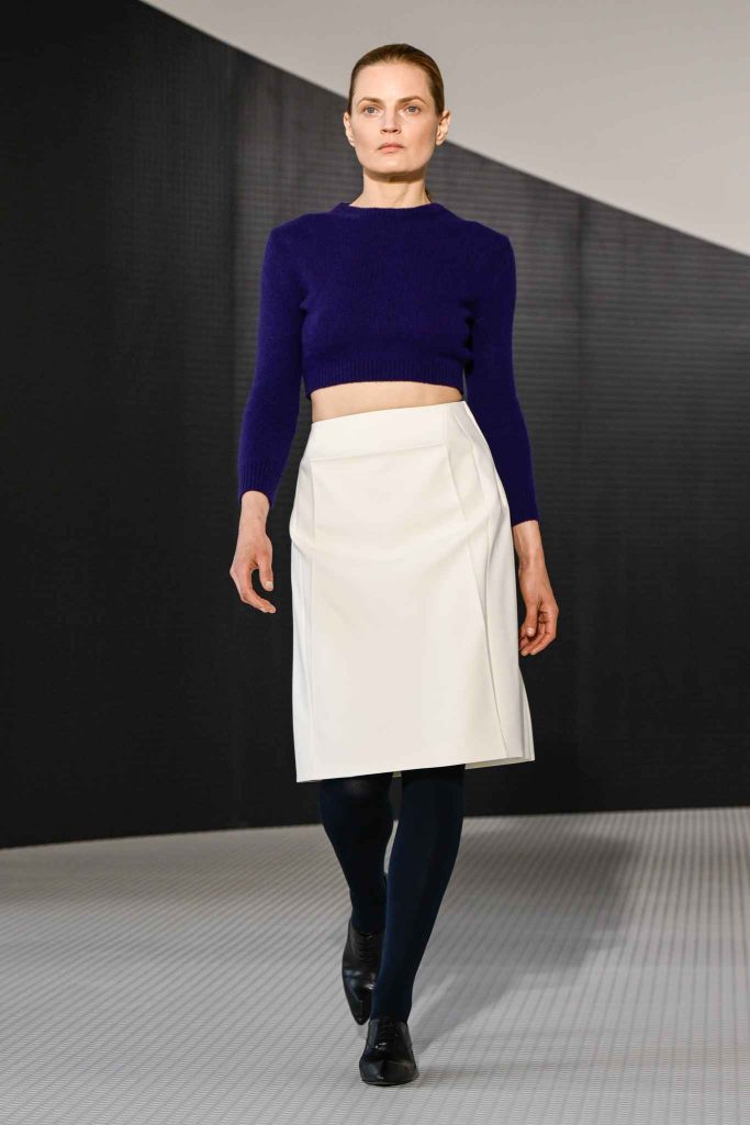 Model in cropped purple knit sweater and white structured knee-length skirt, wearing black tights and oxford shoes, walking on minimalist runway with geometric backdrop.