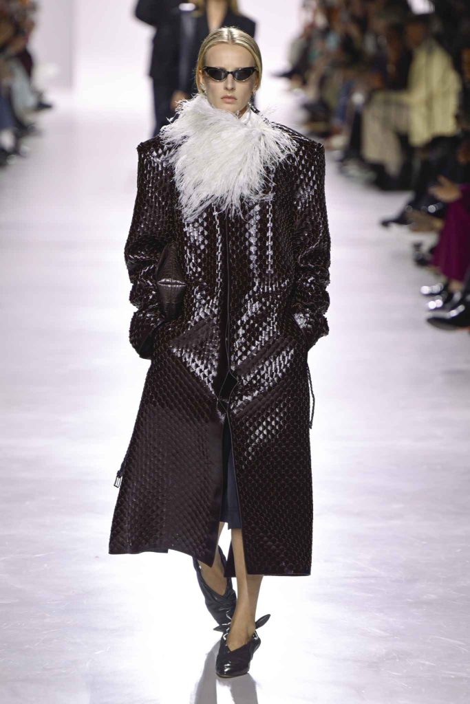 Model in dark textured full-length coat with white feather collar, wearing black cat-eye sunglasses and pointed black shoes, walking on glossy runway with seated audience.