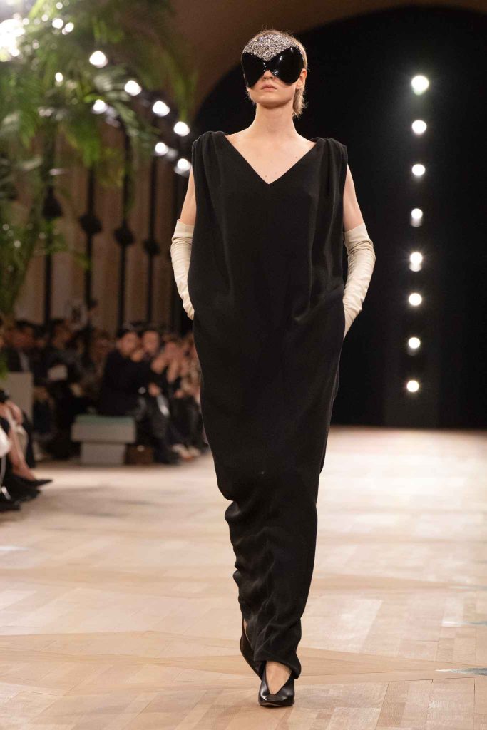 Model in floor-length black column dress with V-neck and cap sleeves, wearing ornate mask and cream opera gloves, walking indoor runway with string lights and foliage backdrop