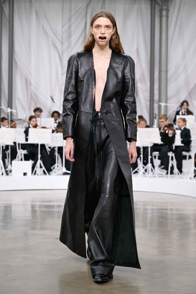 Model in floor-length black leather coat with plunging neckline and wide-leg pants on runway, classical orchestra performing in background