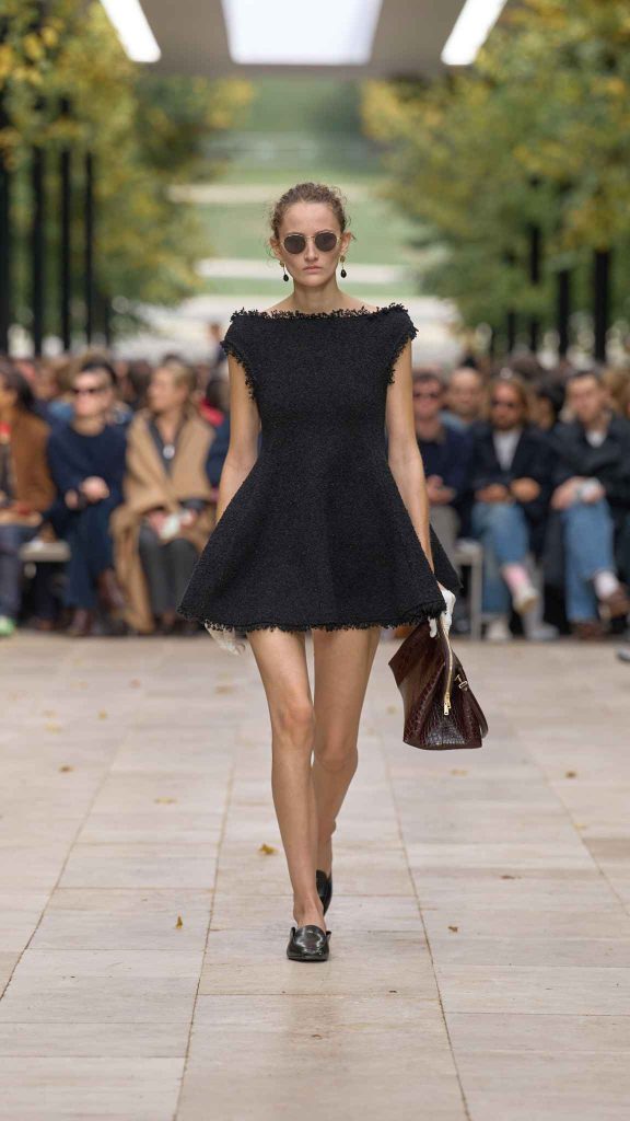 Model in frayed black tweed mini dress with boat neckline and A-line silhouette, wearing sunglasses and carrying brown leather bag on tree-lined outdoor runway