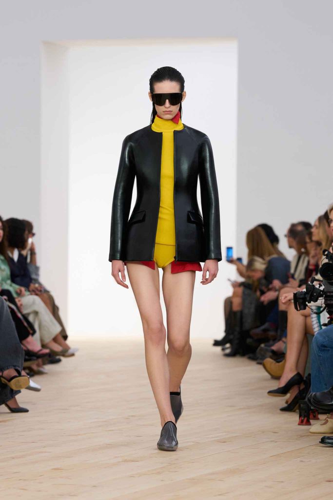 Model in structured black leather jacket over yellow high-neck bodysuit with red shorts, wearing oversized visor sunglasses and gray shoes, walking on minimalist wooden runway with seated audience.