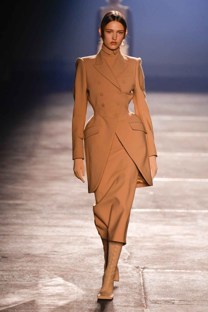 Model wearing camel-colored tailored coat dress with structured shoulders and midi-length skirt on illuminated runway