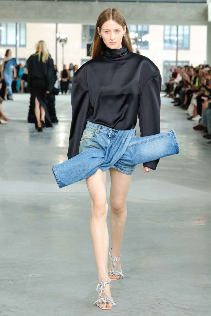 Model wearing oversized black turtleneck with dramatic shoulders and deconstructed denim shorts, paired with silver strappy sandals on concrete runway