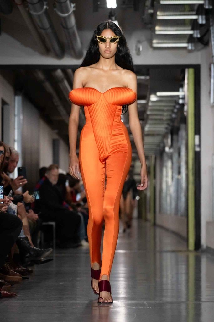 Model wearing structured orange bodysuit with ribbed detailing and gold sunglasses on industrial runway